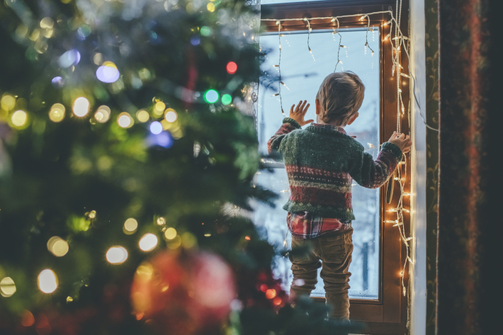 Chłopiec wygląda przez ozdobione świątecznie drzwi balkonu. Obok stoi choinka